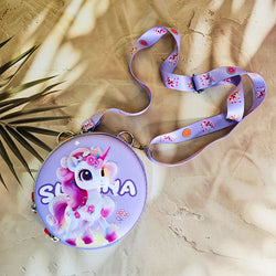 Two colorful, round kids’ sling pouches with attached lanyards are placed on a sandy beige background with a tropical leaf shadow. The left pouch is purple and features an adorable cartoon unicorn with a pink mane, large sparkling eyes, and the name "SHYAMA" written in white. It has a lilac lanyard decorated with candy prints. The right pouch is bright blue with a fierce green cartoon T-Rex dinosaur illustration labeled “DINOSAURS” in bold yellow text. Its lanyard is blue with black dinosaur silhouettes. Bo