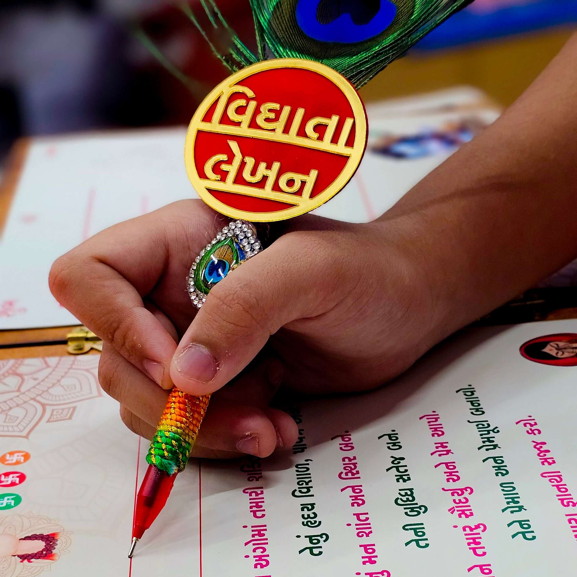 Hand holding a colorful peacock feather pen over a traditional writing board for Chatthi Vidhata Lekhan.