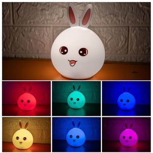 Cute white silicone rabbit night light with pink ears and an adorable face, glowing softly. In the background, two more rabbit lamps glow blue and orange, showcasing the color-changing feature. Perfect as a bedside lamp or nursery decor for kids and adults, providing comforting illumination.www.usmile.com