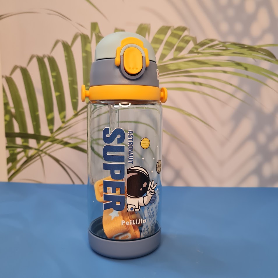 600ml astronaut-themed water bottle for kids featuring a leak-proof design and one-touch flip-open lid. The bottle has a transparent body with space and cartoon graphics, including the word 'SUPER' and an astronaut illustration. The cap is shown in a close-up inset, highlighting the one-key button for opening. The bottle is partially submerged in splashing water, emphasizing its leakproof and durable quality. Available in random colors, ideal for school or travel use