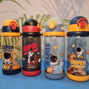 600ml astronaut-themed water bottle for kids featuring a leak-proof design and one-touch flip-open lid. The bottle has a transparent body with space and cartoon graphics, including the word 'SUPER' and an astronaut illustration. The cap is shown in a close-up inset, highlighting the one-key button for opening. The bottle is partially submerged in splashing water, emphasizing its leakproof and durable quality. Available in random colors, ideal for school or travel use