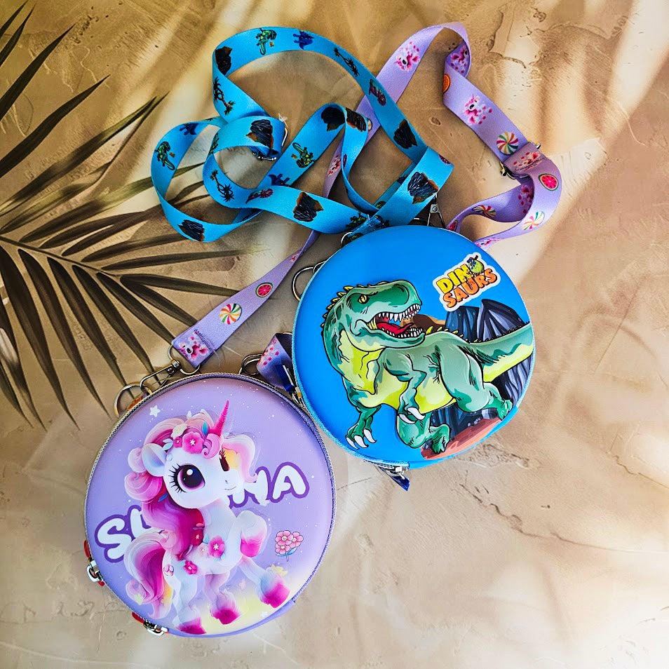 Two colorful, round kids’ sling pouches with attached lanyards are placed on a sandy beige background with a tropical leaf shadow. The left pouch is purple and features an adorable cartoon unicorn with a pink mane, large sparkling eyes, and the name "SHYAMA" written in white. It has a lilac lanyard decorated with candy prints. The right pouch is bright blue with a fierce green cartoon T-Rex dinosaur illustration labeled “DINOSAURS” in bold yellow text. Its lanyard is blue with black dinosaur silhouettes. Bo