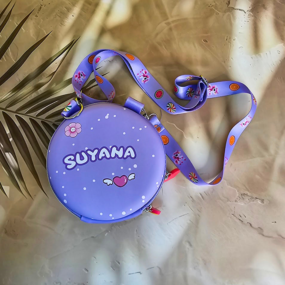 Two colorful, round kids’ sling pouches with attached lanyards are placed on a sandy beige background with a tropical leaf shadow. The left pouch is purple and features an adorable cartoon unicorn with a pink mane, large sparkling eyes, and the name "SHYAMA" written in white. It has a lilac lanyard decorated with candy prints. The right pouch is bright blue with a fierce green cartoon T-Rex dinosaur illustration labeled “DINOSAURS” in bold yellow text. Its lanyard is blue with black dinosaur silhouettes. Bo