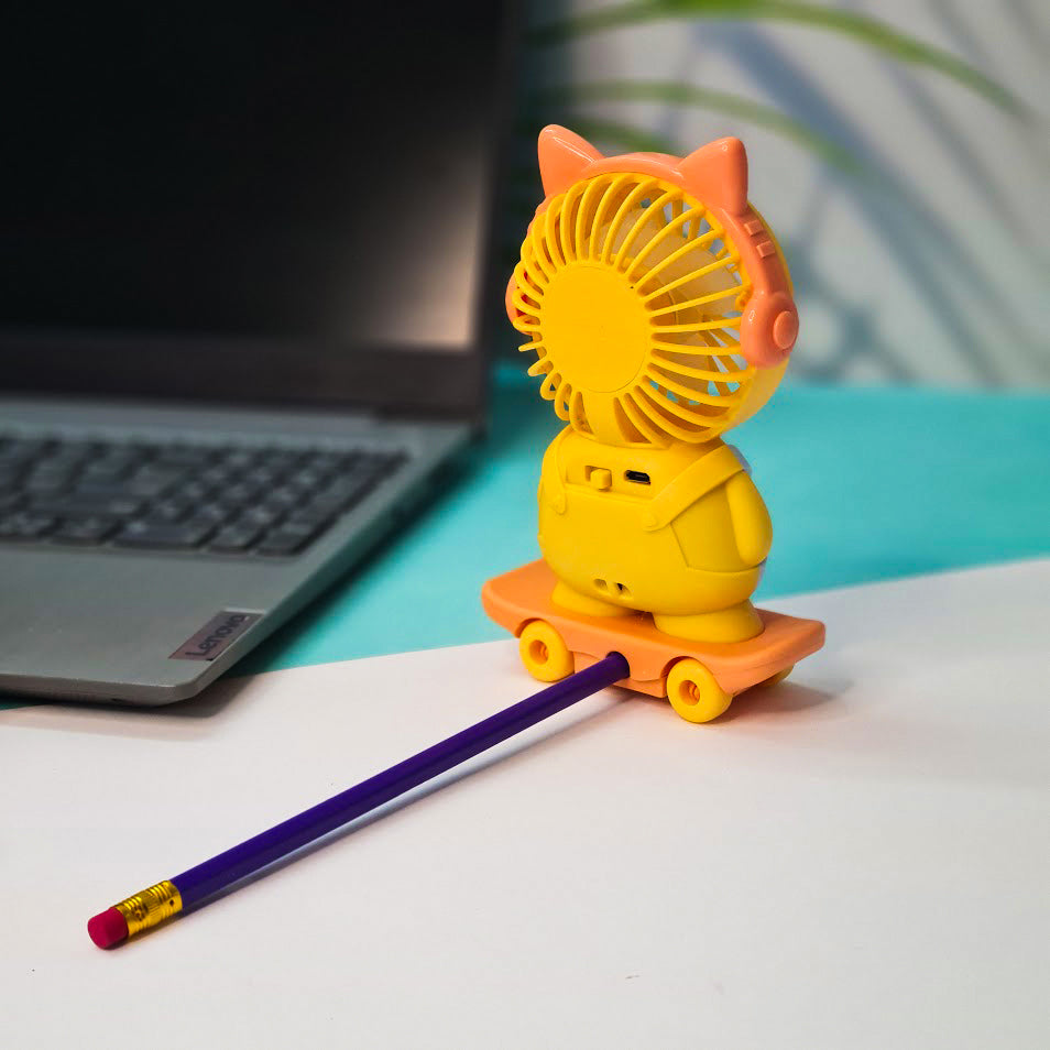 A cute, cartoon-style portable fan shaped like a character standing on a skateboard. The fan's circular blades form the character's face, framed with orange cat ears and orange headphones. The body is yellow with white overalls, giving it a playful and child-friendly appearance. The character stands on an orange toy skateboard with yellow wheels. The fan is placed on a white desk next to a silver Lenovo laptop with a visible keyboard. The background includes a teal surface and a blurred patterned wall with 