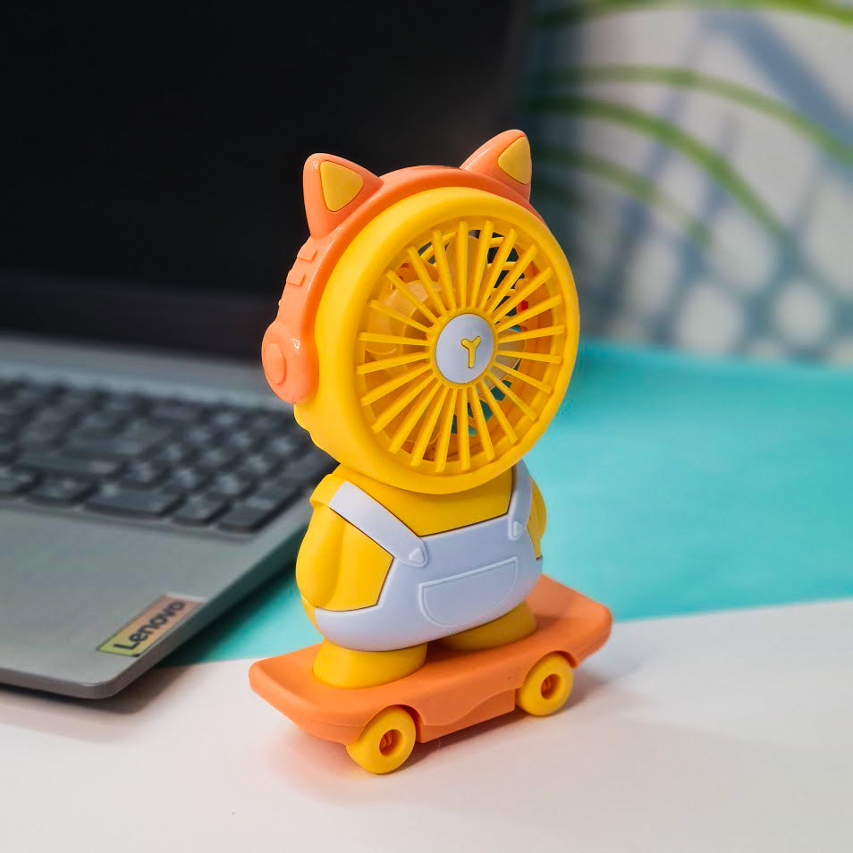 A cute, cartoon-style portable fan shaped like a character standing on a skateboard. The fan's circular blades form the character's face, framed with orange cat ears and orange headphones. The body is yellow with white overalls, giving it a playful and child-friendly appearance. The character stands on an orange toy skateboard with yellow wheels. The fan is placed on a white desk next to a silver Lenovo laptop with a visible keyboard. The background includes a teal surface and a blurred patterned wall with 