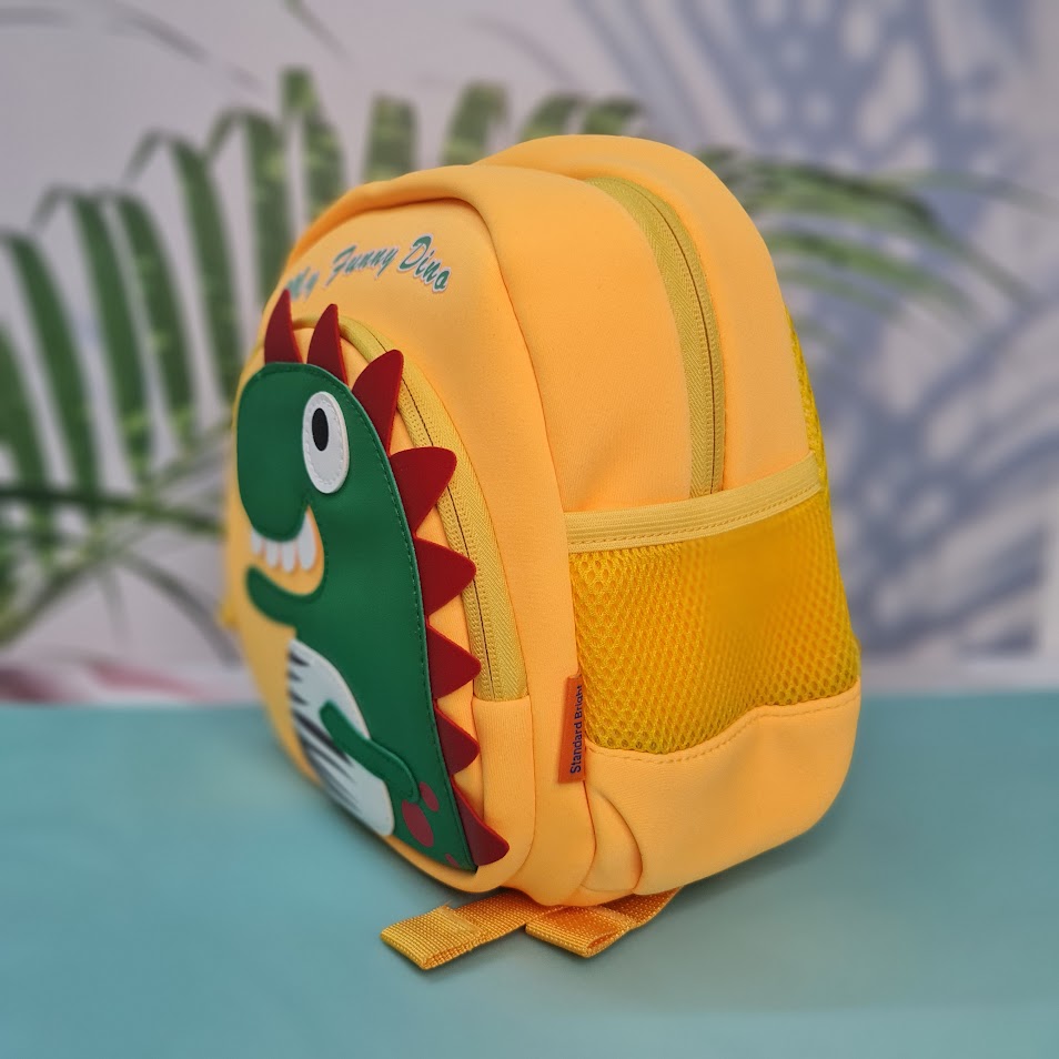 Small, yellow backpack for children featuring a large green dinosaur applique with red spikes and white teeth on the front pocket. Above the dinosaur is embroidered yellow cursive text that reads "My Funny Dino". The backpack has yellow straps and yellow mesh side pockets