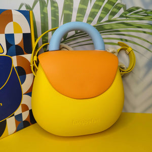 Yellow and orange bag with blue handle on a yellow surface, colorful abstract painting in the background