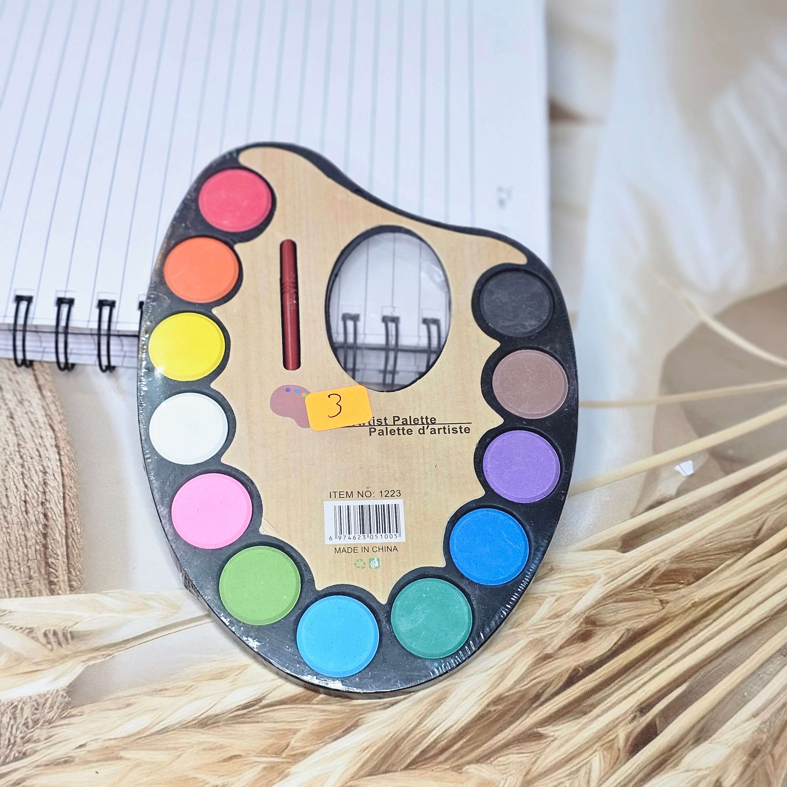 Close-up of a classic-shaped artist's palette containing round pans of assorted watercolors (red, orange, yellow, green, blue, purple, black, white, browns, pink, light blue), with a thin brush in the center, perfect for young painters from Usmile Toys. www.usmile.toys