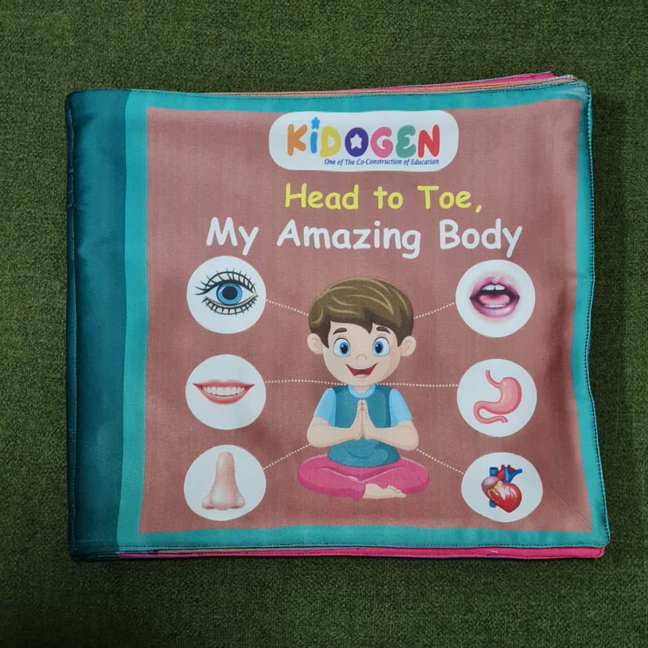 A "My Amazing Body: Head to Toe" fabric book by Kidogen, showing a cartoon child meditating with illustrations of body parts and organs around them.