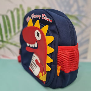 A close-up shot of a navy blue children's backpack. The backpack has a main zippered compartment and a smaller front zippered pocket. The front pocket is adorned with a large, red dinosaur applique with yellow felt spikes, a black and white googly eye, and white felt teeth. The dinosaur also has a white and brown striped detail on its body and yellow circular accents. Above the dinosaur, the phrase "My Funny Dino" is embroidered in a red cursive font. 