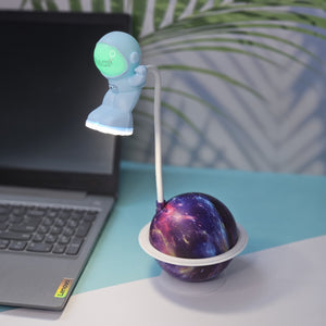 "White astronaut figure desk lamp by Usmile Toys, bending over a glowing purple and blue planet base with a small rocket on top, next to a laptop." www.usmile.toys