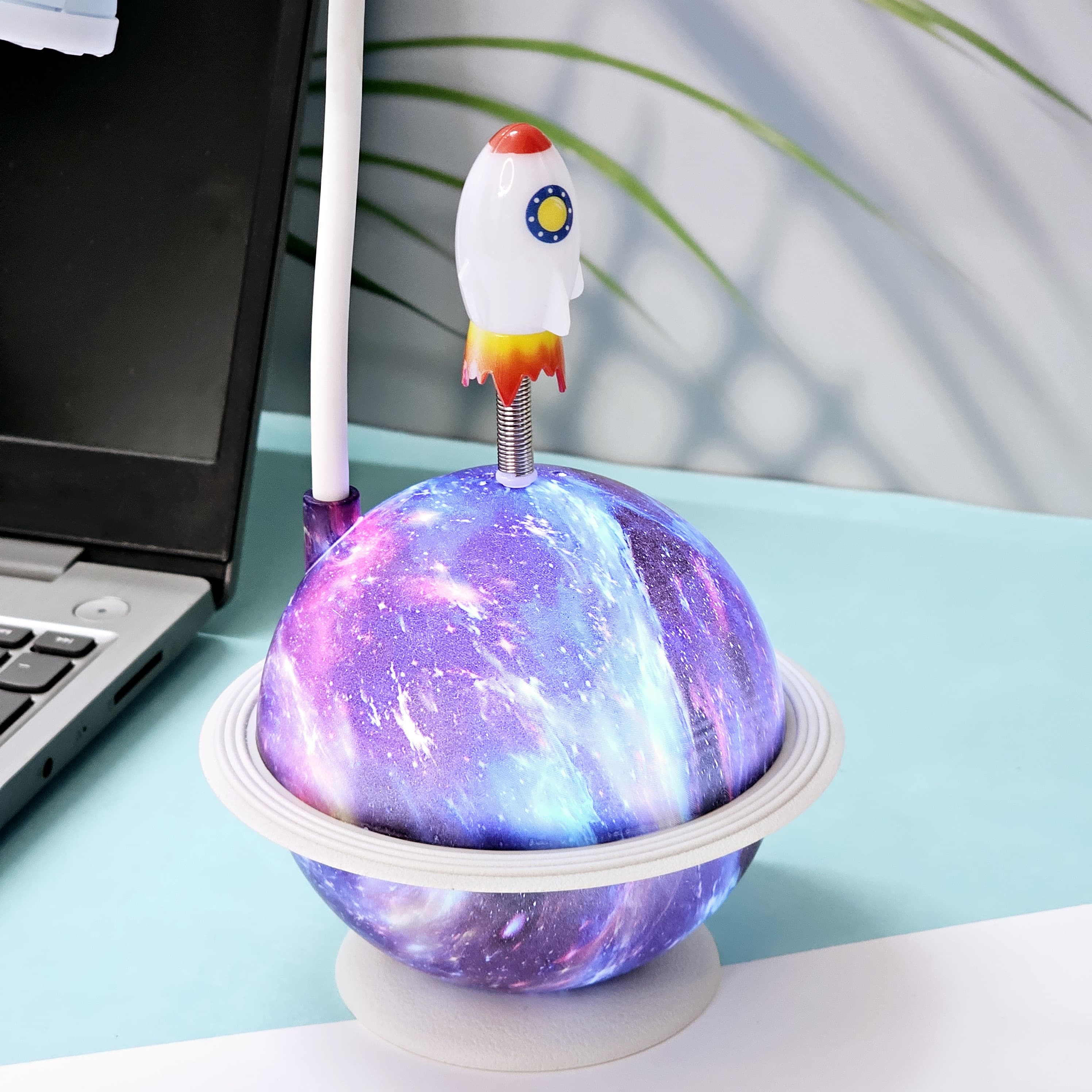 "Close-up of a Usmile Toys creative astronaut lamp, showing its white astronaut figure, flexible arm, and a striking purple and blue glowing planet base with a rocket fin."