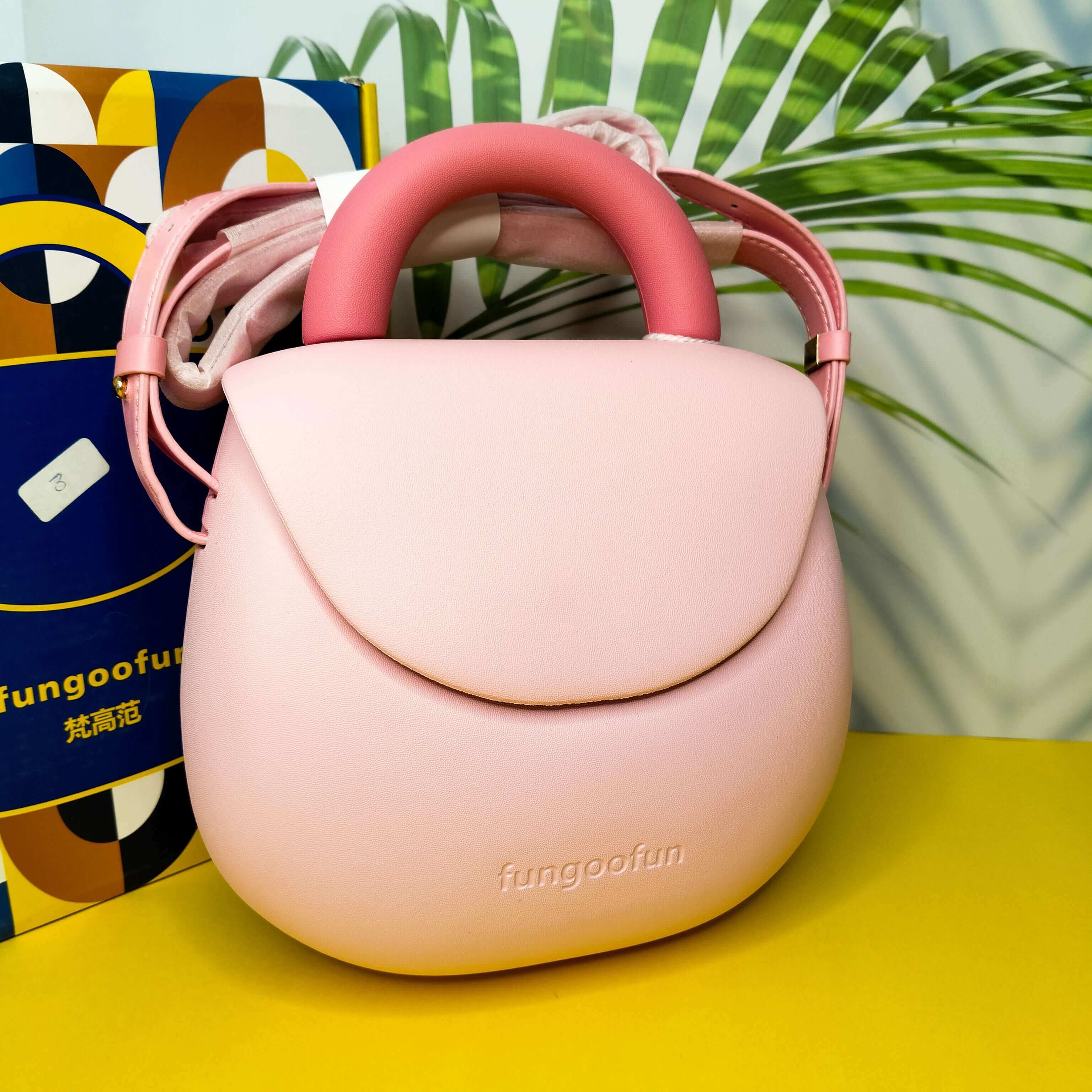 Pink children's handbag with 'fungoofun' branding on a yellow surface with a colorful box and green leaves in the background.