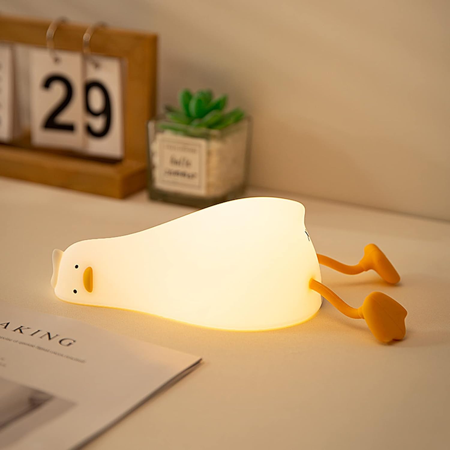dorable glowing white silicone night lamp for kids, shaped like a little duck lying flat with orange feet and beak, resting on a white surface next to a book. Ideal for a calming bedroom ambiance from uSmile.toys." www.usmile,.toys