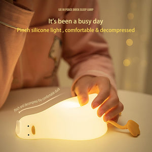 dorable glowing white silicone night lamp for kids, shaped like a little duck lying flat with orange feet and beak, resting on a white surface next to a book. Ideal for a calming bedroom ambiance from uSmile.toys." www,usmile.toys