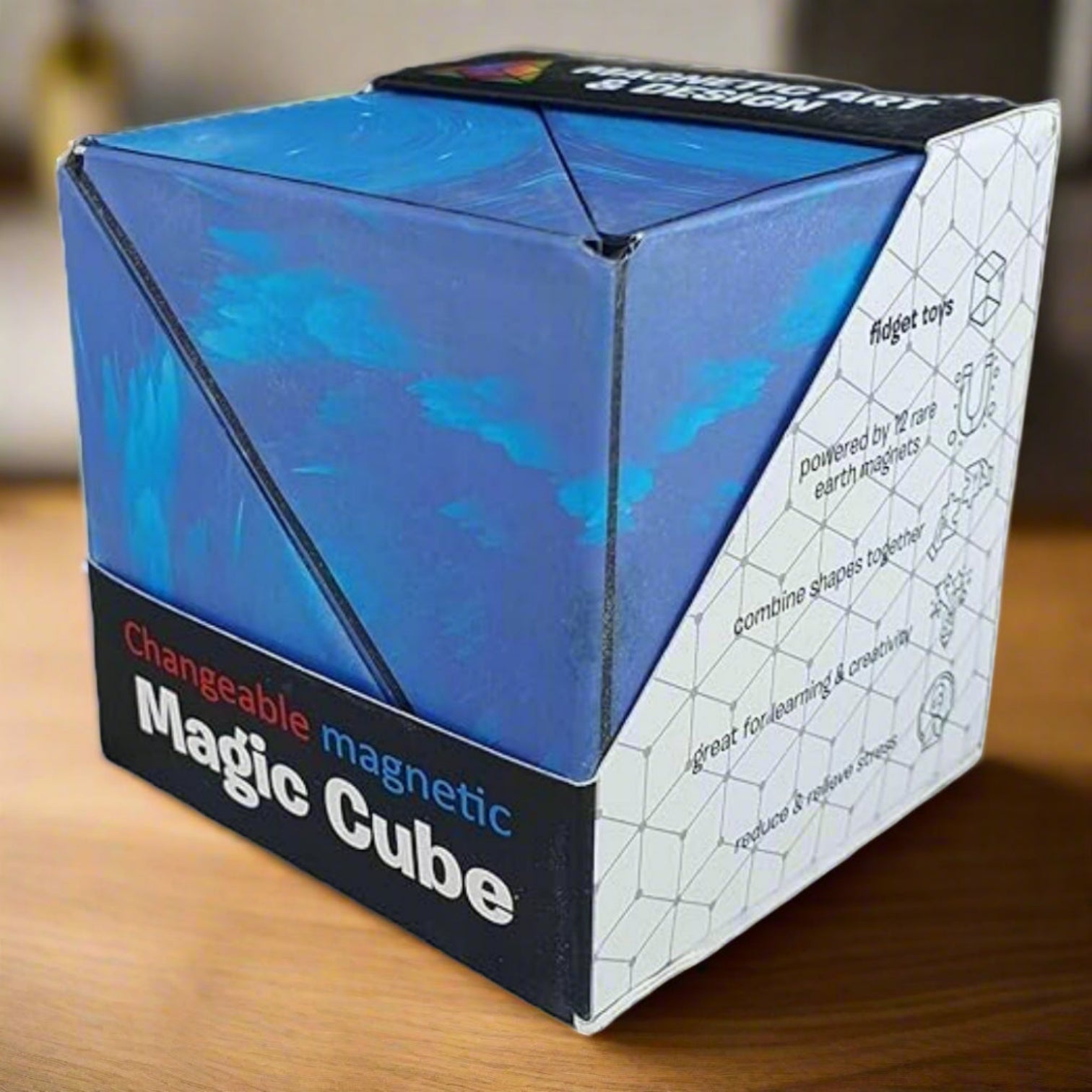 A photo of a boxed product called the “Changeable Magnetic Magic Cube.” The cube is displayed inside a unique packaging that emphasizes its geometric, triangular folding structure. The visible part of the cube has a vibrant, abstract blue design resembling swirling ocean or sky patterns. The box features various marketing phrases. The front side displays the name in bold white font: “Magic Cube,” with "Changeable" in red and "magnetic" in blue, highlighting its changeable and magnetic nature. The right side