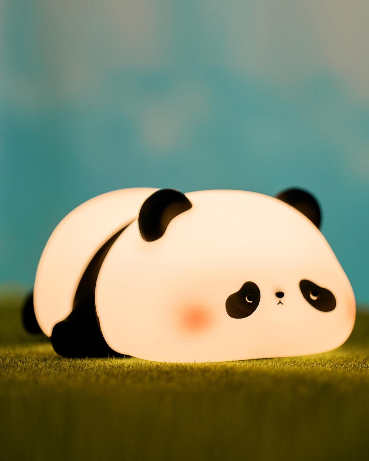 Adorable glowing white and black silicone night lamp for kids, shaped like a cute lying flat panda, resting on green grass against a blue sky. Perfect for a calming bedroom ambiance from uSmile.toys." www.usmile.toys