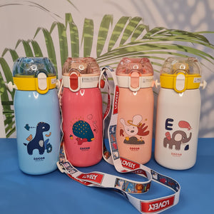 Four adorable and colorful children's water bottles are displayed against a backdrop of blurred green and white palm leaf decorations. Each bottle is made of stainless steel and features a double-walled, vacuum-insulated design to keep drinks cold or warm. They have a rounded, cylindrical shape with a slightly tapered neck leading to a flip-top lid with a straw. The lids are translucent plastic in blue, light pink, coral, and clear yellow, each with a contrasting yellow or light pink band around the base. 